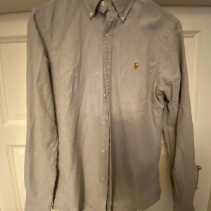 Ralph lauren skjorta - Klassisk grå skjorta från Ralph Lauren med button-down krage och broderad logga på bröstet. Skjortan är långärmad och har knappar framtill. Tillverkad i mjuk bomull som känns skön mot huden. Perfekt för en clean och stilren look.