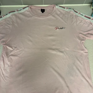 Ljusrosa t-shirt från Prolific - Ljusrosa t-shirt från Prolific med korta ärmar och logga broderad på bröstet. Snygga band med Prolific-tryck och ränder i svart, rött och vitt längs axlarna. Skön passform och perfekt för en chill stil. 