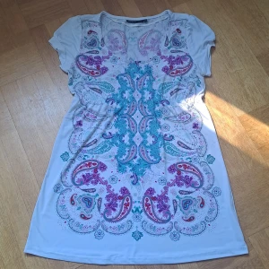 Vit kortklänning med paisleymönster - Kortklänning från äldre KappAhl i vitt med färgglatt paisleymönster i turkos, lila och rött. Klänningen har korta ärmar och rund halsringning. Mjuk och lätt i materialet, perfekt för varma dagar. 🩷