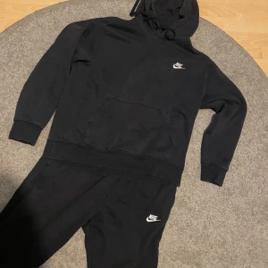 Svart Nike hoodie med huva och byxor - Svart hoodie från Nike med klassisk vit logga på bröstet. Tröjan har huva med snörning, stor magficka och långa ärmar med muddar. Perfekt för chill dagar eller när du vill ha en clean streetstyle-look.