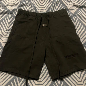 Svarta shorts Essentials Fear of God - Svarta shorts från Essentials Fear of God med broderad logga på vänster ben. Modellen har hög midja, elastisk midja med snörning och två sidofickor. Tillverkade i mjuk bomull, perfekta för chill dagar eller streetwear-stil.