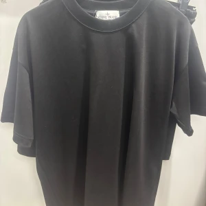 Svart Stone Island t-shirt med patch Set - Svart t-shirt från Stone Island med klassisk rund hals och korta ärmar. På ena ärmen sitter den ikoniska Stone Island-patchen med gult broderi och svarta knappar samt Shorten. T-shirten& Shortsen är i bomull och har en clean, loose passform som ger en chill vibe.