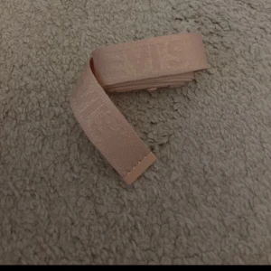 Rosa Levi's tygbälte med spänne - Säljer ett beige tygbälte från Levi's med diskret logga invävd i bandet. Bältet har ett matchande beige spänne i plast och är smalt och flexibelt, perfekt för att ge en clean look till jeans eller byxor.