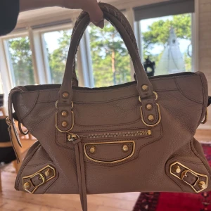 Beige Balenciaga handväska i skinn - Säljer en beige handväska från Balenciaga i äkta skinn med guldiga metalldetaljer och dragkedja framtill. Väskan har två flätade handtag och ikoniska nitar. Perfekt storlek för det viktigaste och en snygg detalj till din outfit.