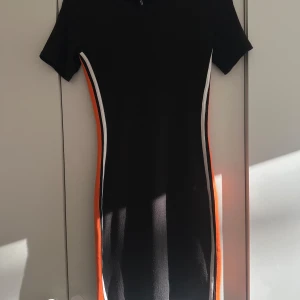 Svart klänning med ränder - Svart klänning med sportiga vita och orange ränder längs sidorna. Tight passform och kortärmad modell som ger en cool och modern vibe. Perfekt för dig som gillar en enkel men ändå färgstark look