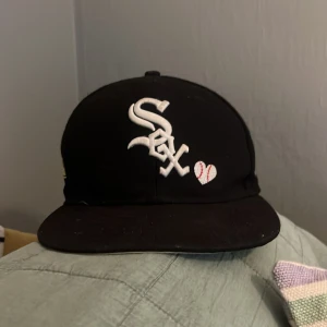 Svart White Sox keps New Era  - Snygg svart keps från New Era 59FIFTY med White Sox-logga framtill och broderat hjärta. Baksidan har texten 'Love & Hustle = Game' och MLB-logga. Klassisk rak skärm och broderade detaljer. Perfekt för dig som gillar streetstyle och sport. Priset är inte hugget i sten!😁