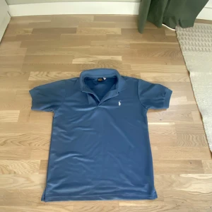 Blå pikétröja från Polo Ralph Lauren - Säljer en klassisk blå pikétröja från Polo Ralph Lauren i storlek S. Tröjan har korta ärmar, krage och den ikoniska broderade loggan på bröstet. Perfekt för en clean och stilren look. Materialet känns som bomull och är riktigt skönt mot huden.