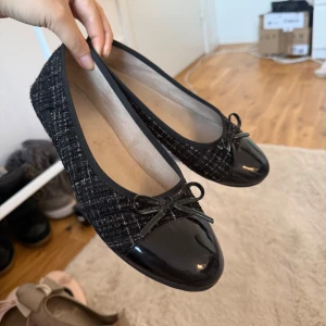 Svarta ballerinaskor med lackad tå - Stilrena svarta ballerinaskor med glansig lackad tå och söt rosett framtill. Ovandelen har ett diskret rutigt tygmönster som ger en lyxig känsla. Perfekta för dig som gillar klassisk och chic stil.