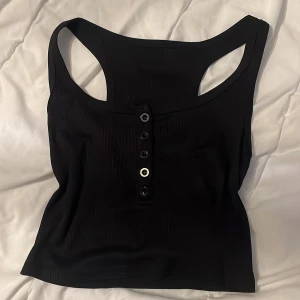 Svart ribbad linnetopp med knappar - Säljer en svart ribbad linnetopp med snygga knappar framtill. Toppen har brottarrygg och är croppad, perfekt till jeans eller kjol. Materialet är stretchigt och sitter skönt mot kroppen. Enkel men ändå cool detalj med knapparna framtill.