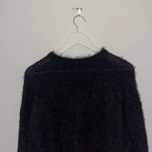 Svart fluffig stickad tröja Tiffosi - Mysig svart stickad tröja från Tiffosi i storlek M. Tröjan har en fluffig och lite lurvig textur som ger en cool och trendig look. Den har rund halsringning och långa ärmar, perfekt för kyliga dagar.