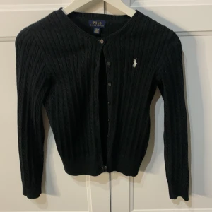 Svart kabelstickad kofta Polo Ralph Lauren - Kabelstickad svart kofta från Polo Ralph Lauren med klassisk rund hals och knappar framtill. Storlek 12-14 år använd några ggr.