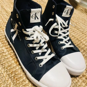 Marinblå höga sneakers Calvin Klein - Snygga marinblå höga sneakers från Calvin Klein Jeans med vit tå och vita snören. Skorna har tydlig CK-logga på sidan och är gjorda i canvasmaterial. Klassisk rund tå och platt sula, perfekt för en clean streetstyle-look. Använda två gånger.