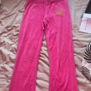 Juicy Couture rosa mjukisbyxor XL - Rosa mjukisbyxor från Juicy Couture med broderad guldfärgad krona och texten 'preppy royalty JUICY' framtill. Byxorna har bred resår i midjan, snörning och är tillverkade i mjuk velour. Perfekta för en chill och trendig look.