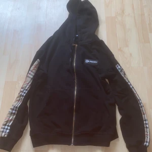 Svart Burberry hoodie med rutig detalj - Svart hoodie från Burberry med dragkedja och huva. Ärmarna har klassiska Burberry-rutiga paneler med stor logga längs sidorna. Jackan har fickor framtill och en patch med Burberry-logga på bröstet. Perfekt för dig som vill sticka ut med en ikonisk streetstyle.