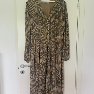 Zebramönstrad långklänning från Adoore - Snygg långklänning från Adoore med zebramönster i beige och svart. Klänningen har v-ringning, långa ärmar och knappar framtill. Perfekt för dig som vill sticka ut med en cool och trendig look. Använd endast vid fåtal tillfällen. 