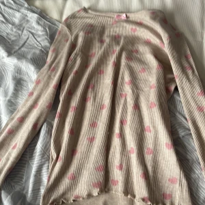 Beige ribbad långärmad tröja med hjärtan - Supersöt beige ribbad långärmad tröja med rosa hjärtan över hela och en liten rosa rosett vid halsen. Tröjan har volangkant nertill och vid ärmsluten, vilket ger en gullig vibe. Perfekt för dig som gillar mjuka färger och detaljer.
