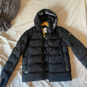 Svart Moncler dunjacka med huva - Säljer en svart dunjacka från Moncler med fet logga på ärmen och tryckt Moncler-text på insidan av huvan. Jackan har dragkedjor på fickorna och är quiltad för extra värme. Perfekt för kalla dagar och har en clean, modern look.