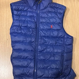 Blå dunväst från Polo Ralph Lauren - Säljer en blå dunväst från Polo Ralph Lauren med quiltad design och dragkedja framtill. Västen har hög krage, två sidofickor med dragkedja och den klassiska röda Polo-loggan broderad på bröstet. Perfekt för lager-på-lager under kyliga dagar.