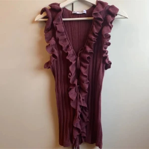 Vinröd ribbad volangtopp - Supersnygg vinröd ärmlös topp med djup v-ringning och volangdetaljer längs kanterna. Toppen är ribbad och har knyt framtill för extra stilpoäng. Perfekt för dig som vill sticka ut med en trendig och feminin look.