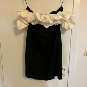 Svart kortklänning med volang - Supersnygg svart kortklänning med stora vita volanger upptill. Klänningen har en trendig offshoulder-look och passar perfekt när du vill sticka ut. Materialet känns lätt och bekvämt, och volangdetaljen ger en cool statement-effekt. Aldrig använd. 
