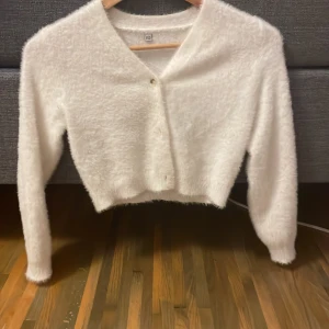 Vit fluffig kofta med knapp - Supermjuk och fluffig vit kofta med croppad passform och v-ringning. Koftan har 3 enkla knappar framtill och långa ärmar. Perfekt för lager-på-lager och ger en mysig vibe till din outfit.