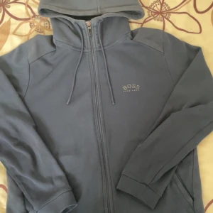 Blå hoodie från Hugo Boss - Snygg blå hoodie från Hugo Boss med dragkedja hela vägen och huva med snörning. Tröjan har diskret BOSS-logga på bröstet och är tillverkad i mjukt material, perfekt för chill dagar. Långärmad och med ribbade muddar för extra komfort.