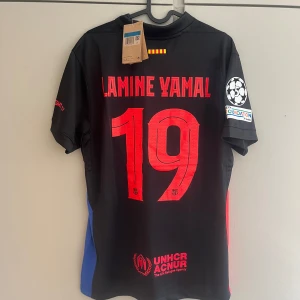 FC Barcelona Lamine Yamal #19 Nike tröja - Snygg svart FC Barcelona fotbollströja från Nike med Lamine Yamal #19 på ryggen. Tröjan har röda och blå detaljer på sidorna, röda tryck och klubbmärke på bröstet. Champions League-märke på ärmen och sponsorer som Spotify och UNHCR. Tillverkad i lätt och ventilerande polyester.