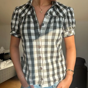 Rutig kortärmad skjorta med puffärm - Snygg skjorta med svart och vit rutigt mönster, korta puffärmar och knappar framtill. Skjortan har klassisk krage och är gjord i ett lätt bomullstyg, perfekt till jeans eller kjol. Passformen är normal och den har en fräsch, ungdomlig vibe.