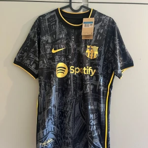 FC Barcelona svart matchtröja Nike - Snygg FC Barcelona fotbollströja i svart med guldiga detaljer och tryck från Nike. Tröjan har korta ärmar, gul logga och sponsortryck, samt ett unikt grafiskt mönster över hela tyget. Tillverkad i lätt och ventilerande polyester, perfekt för match eller träning.