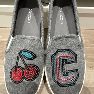 Grå loafers med körsbär och bokstav - Grå loafers från H&M Divided i filtad syntet med broderade detaljer: ett körsbär i rött och blått på ena skon och ett stort C i rosa, blått och svart på den andra. Skorna har vit sula och är perfekta för en lekfull och unik stil.