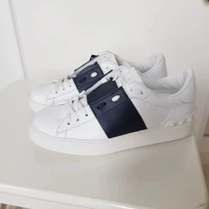 Valentino Open sneakers vit/marinblå - Snygga Valentino Open sneakers i vitt skinn med en bred marinblå rem över mitten. Klassisk rund tå, platt sula och diskreta nitar baktill. Perfekta för dig som gillar stilrena och exklusiva sneakers med en twist.