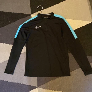 Nike svart träningströja med blå detaljer - Svart långärmad träningströja från Nike med ljusblå ränder längs axlarna och halv dragkedja framtill. Perfekt för fotboll eller gymmet, med sportig look och bekväm passform. Swoosh-logga på bröstet ger extra stilpoäng.