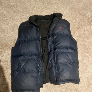 Mörkblå dunväst från Polo Ralph Lauren - Snygg mörkblå dunväst från Polo Ralph Lauren med hög krage och klassisk röd logga på bröstet. Västen har tryckknappar, dragkedja och två stora fickor framtill. Perfekt att slänga över en hoodie eller t-shirt för en chill look. Storlek M. 