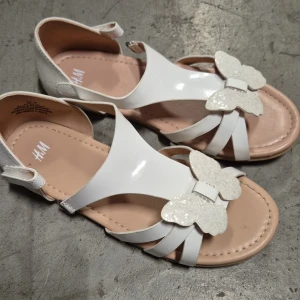 Sandaler - Söta vita sandaler från H&M med glittriga fjärilar framtill. Skorna har öppen tå, remmar och justerbar rem runt hälen. Insidan är ljusbeige och baksidan har en lätt skimrande yta. Perfekta för varma dagar och somriga outfits.