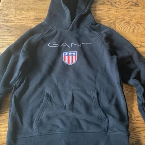 Svart hoodie från GANT med logga - Svart hoodie från GANT med broderad logga och amerikanskt sköldmärke på bröstet. Klassisk känguruficka framtill och huva. Perfekt för dig som gillar en sportig och stilren look.