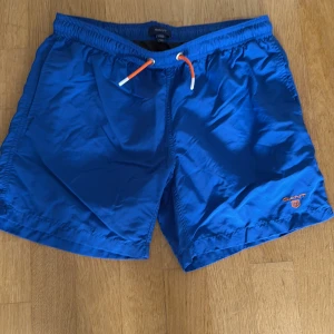 Blå badshorts från Gant med snörning - Snygga blå badshorts från Gant med orange snörning i midjan och broderad logga på benet. Shortsen har elastisk midja och är perfekta för stranden eller poolen. Lätta och snabbtorkande material som känns fräscht hela sommaren.