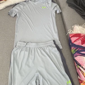 Ljusgrå tränings-tshirt från Under Armour - Ljusgrå t-shirt från Under Armour med kort ärm och diskret neongrön logga på bröstet. Tillverkad i ett lätt och svalt polyestermaterial som passar perfekt för träning. Enkel och stilren design med sportig känsla.