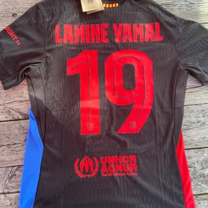 FC Barcelona Lamine Yamal Champions League tröja - Säljer en svart FC Barcelona Champions League-tröja med Lamine Yamal och nummer 19 på ryggen. Tröjan har röda och blå detaljer på sidorna, röda tryck och klubbmärke på bröstet. Nike-logga, Spotify-sponsor och UEFA Foundation-märke på ärmen. Tillverkad i ventilerande funktionsmaterial.