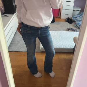 Blå bootcut jeans från H&M - Säljer ett par blå bootcut jeans från H&M med låg midja och justerbar midja. Jeansen har dragkedjedetaljer framtill och klassiska bakfickor. Materialet är en mix av bomull, polyester och elastan för en skön passform.