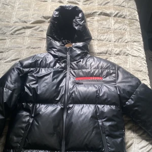 Svart pufferjacka från Prada - Säljer en svart pufferjacka från Prada med glansig finish och huva. Jackan har dragkedja framtill, två sidofickor och en bröstficka med röd detalj och Prada-logga. Perfekt för kalla dagar och riktigt snygg streetstil.