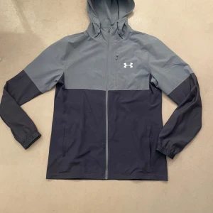 Grå och svart vindjacka Under Armour - Snygg tvåfärgad vindjacka från Under Armour i grått och svart. Jackan har huva, dragkedja framtill och diskret logga på bröstet. Perfekt för dig som gillar sportig stil och vill ha något lätt och smidigt till vardagen. Helt ny använt 1 gång