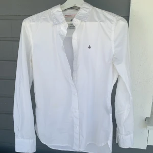 Vit skjorta med ankare från Soft, Layer & Strength - Klassisk vit skjorta med liten broderad ankare på bröstet från Soft, Layer & Strength. Skjortan har lång ärm, krage och knäppning framtill. Perfekt till många olika outfits och enkel att styla.