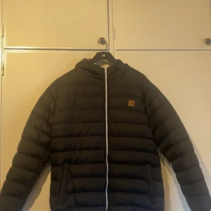 Svart pufferjacka från Urban Classics - Säljer en svart pufferjacka från Urban Classics i storlek XXL. Jackan har huva, dragkedja framtill och två sidofickor. Klassisk quiltad design med en liten patch på bröstet. Perfekt för kyliga dagar. Den är endast använd 2 gånger, priset kan diskuteras!
