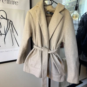 Beige kappa med bälte från Zara - Stilren beige kappa från Zara med klassisk krage och knytskärp i midjan. Jackan har långa ärmar och två fickor framtill. Perfekt för dig som vill ha en tidlös och elegant look. Jackan är tyvärr ganska nopprig över hela fram och baksida 
