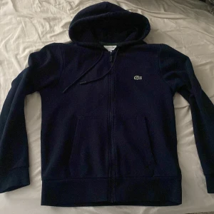 Mörkblå hoodie från Lacoste - Säljer en mörkblå zip hoodie från Lacoste med dragkedja och huva. Klassisk krokodillogga på bröstet och två fickor framtill. Inga skador, nästan oanvänd pga fel storlek. Tveka inte på att skriva vid funderingar😁