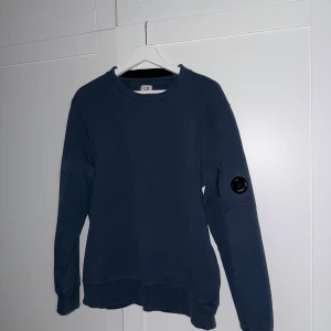 Blå sweatshirt från C.P. Company - Säljer en stilren blå sweatshirt från C.P. Company med rund hals och lång ärm. Tröjan har en ikonisk linsdetalj på ena ärmen och är tillverkad i mjukt bomullsmaterial. Perfekt för en avslappnad och trendig look.
