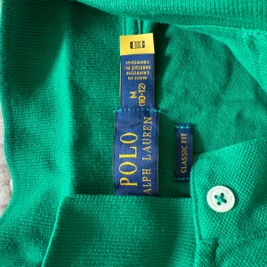 Grön pikétröja från Polo Ralph Lauren - Säljer en klassisk grön pikétröja från Polo Ralph Lauren i storlek M. Tröjan har lång ärm, krage och knäppning framtill samt den ikoniska lilla loggan broderad på bröstet. Perfekt för en stilren och sportig look.