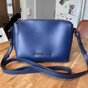 Blå axelväska från Armani Jeans - Snygg blå axelväska från Armani Jeans med stilren design och justerbar axelrem. Väskan har dragkedja upptill och ett mönstrat innerfoder. Perfekt för att bära det viktigaste med stil.Helt ny