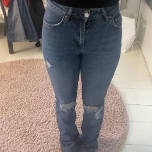 Blå midways jeans med slitningar - Säljer ett par blå högmidjade jeans med utsvängdning och snygga slitningar på båda knäna. Jeansen har klassisk femficksdesign och stängs med knapp och dragkedja. Perfekta för en avslappnad och trendig look.