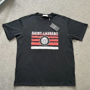 Saint Laurent T-shirt  - Svart t-shirt från Saint Laurent med vitt och rött tryck framtill. T-shirten har rund halsringning och korta ärmar. Materialet är mjukt och skönt, perfekt för en avslappnad stil. Storlek Large men sitter mer som medium. 10/10 skick då tröjan aldrig är använd och har lappen kvar som du kan se på bilden! Bara höra av dig vid frågor/ funderingar! 